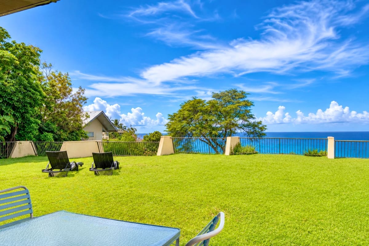 A grassy outdoor area with a table and chairs, a building, and a view of the ocean.