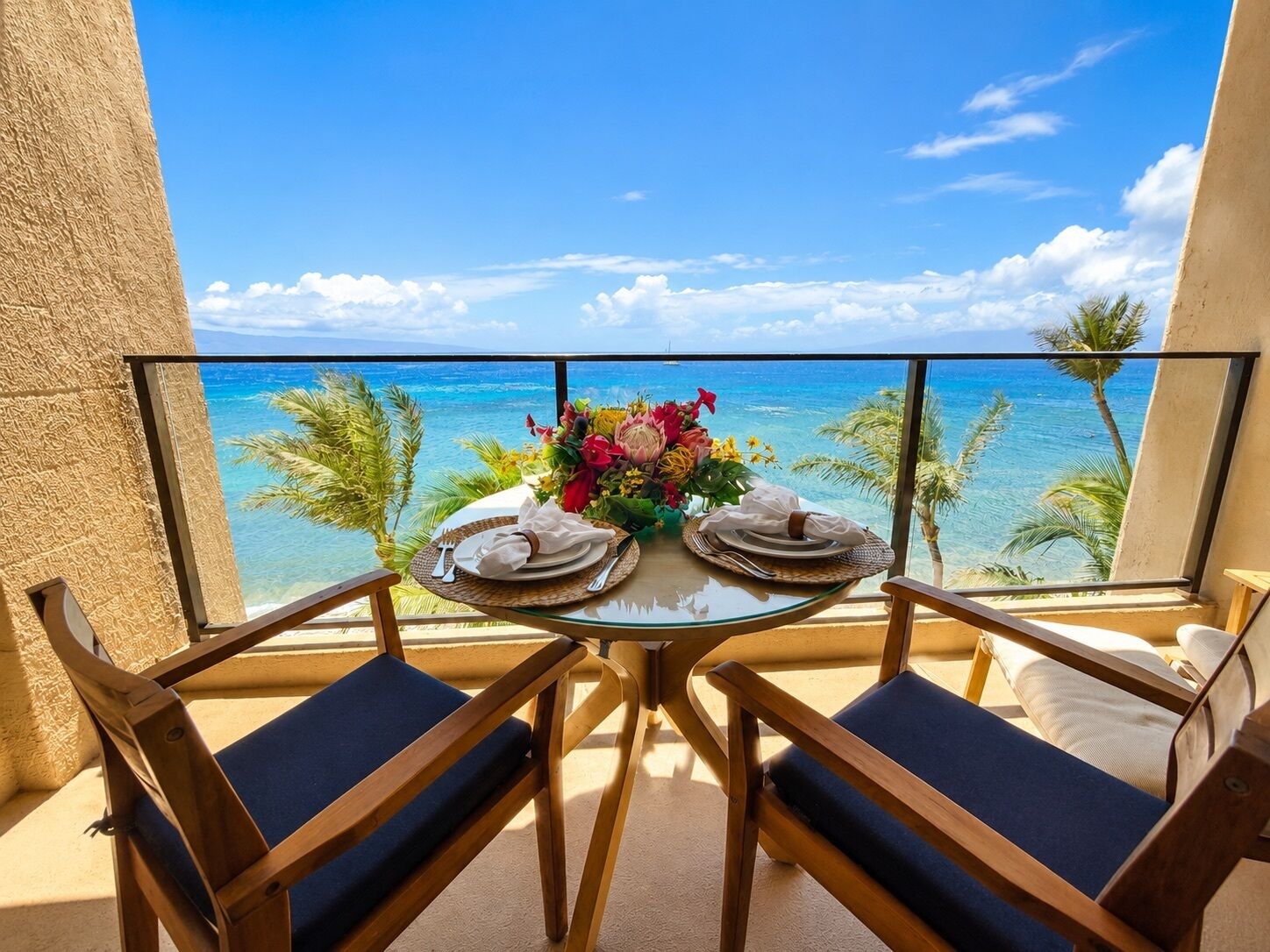A balcony with a table set for two, overlooking a clear blue sea and palm trees.