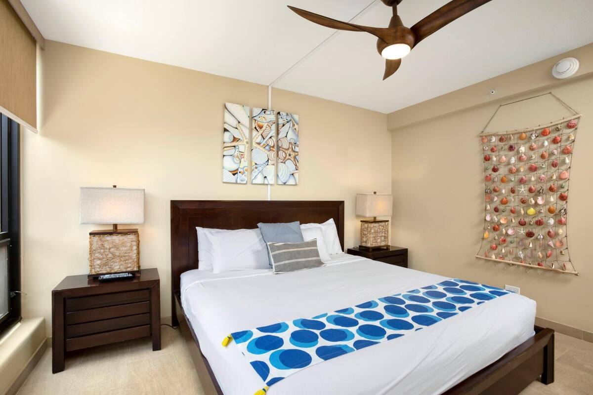 A hotel room with a bed, two bedside tables with lamps, a ceiling fan, a wall with a decorative shell wall hanging, and a window with blinds.