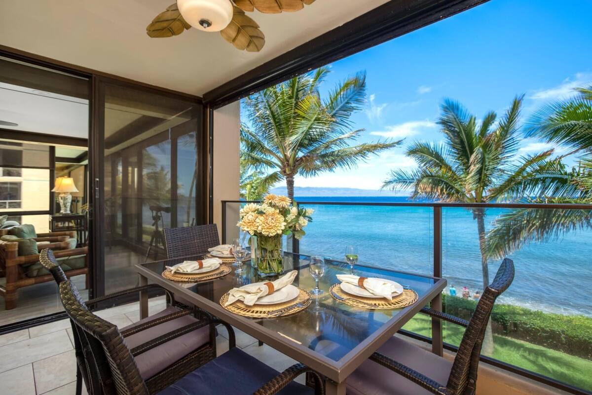 A dining area with a glass table and wicker chairs, overlooking a clear blue sea with palm trees.