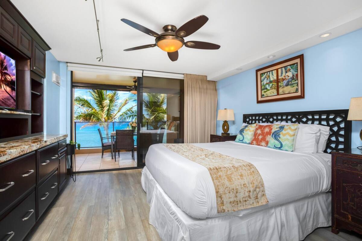 A hotel room with a bed, a ceiling fan, a painting, and a view of palm trees.