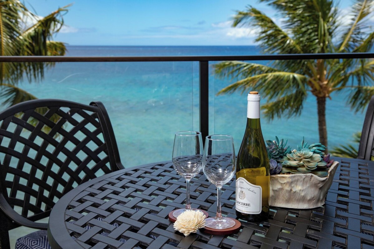 A table with a bottle of wine, two glasses, and a flower centerpiece, overlooking a tropical beach with palm trees.