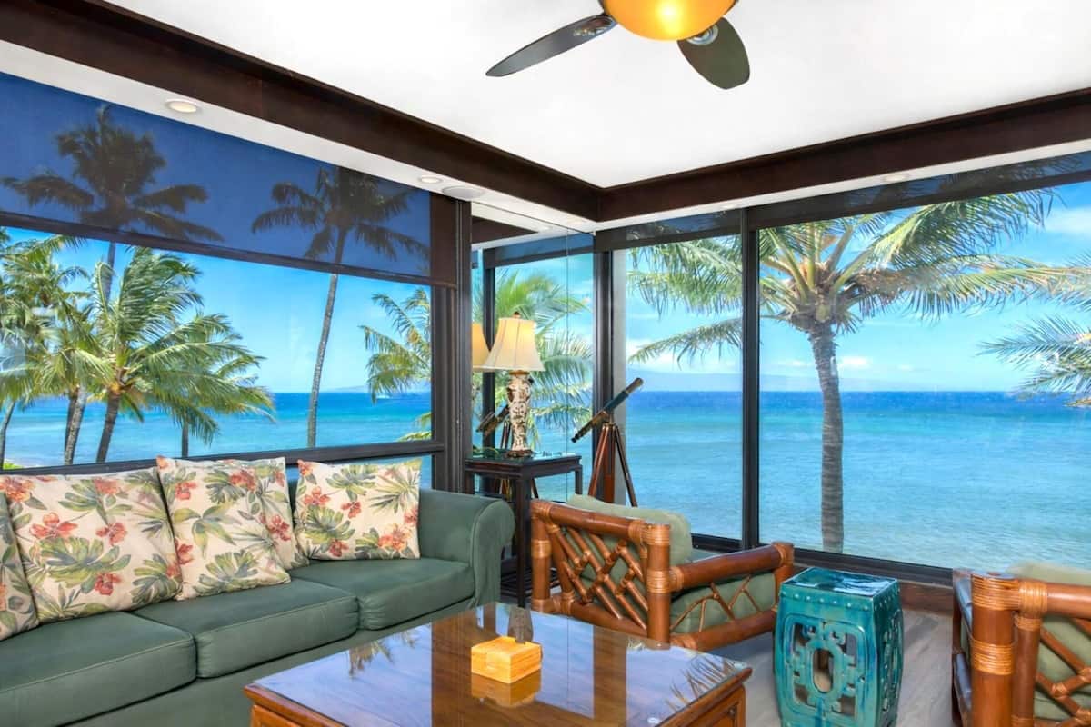 A room with a green sofa, a wooden coffee table, and a view of the ocean through large windows.