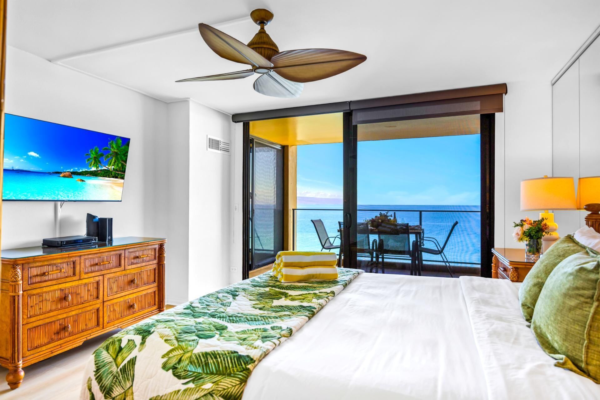 A hotel room with a bed, a TV, a wooden dresser, a balcony with a table and chairs, and a view of the sea.