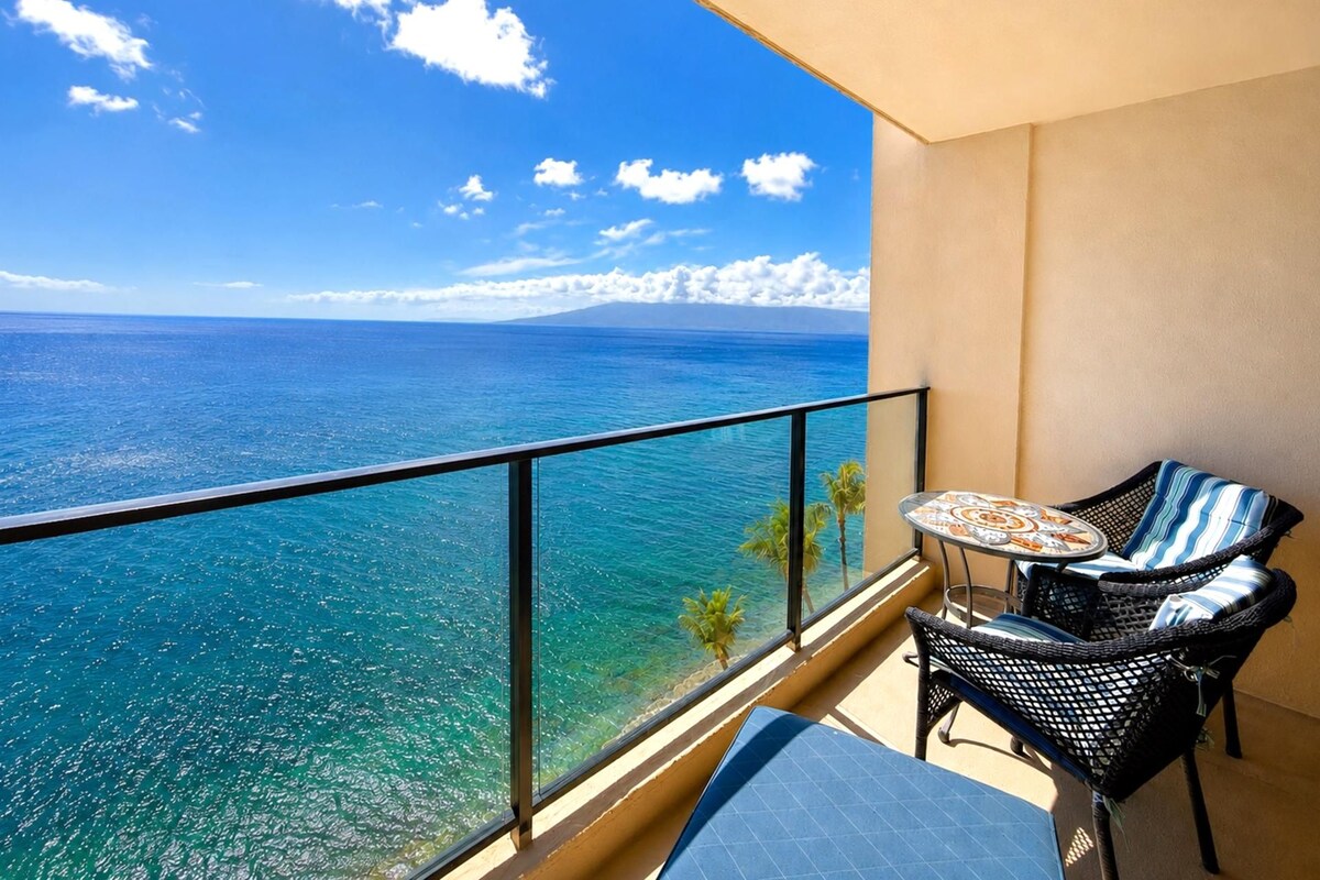 A balcony with a clear view of the ocean, a small table, two wicker chairs, and a single palm tree.