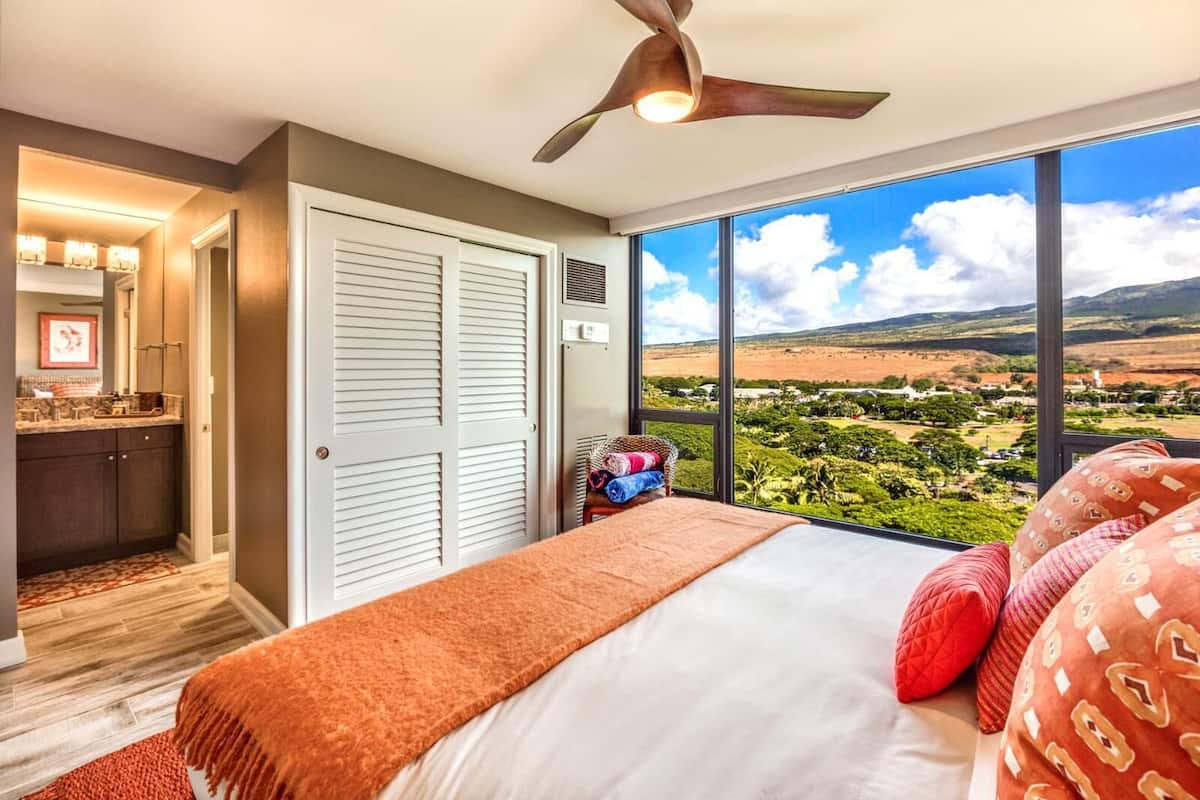 A bedroom with a bed, a ceiling fan, a large window with a view of a landscape, and a bathroom with a vanity and mirror.