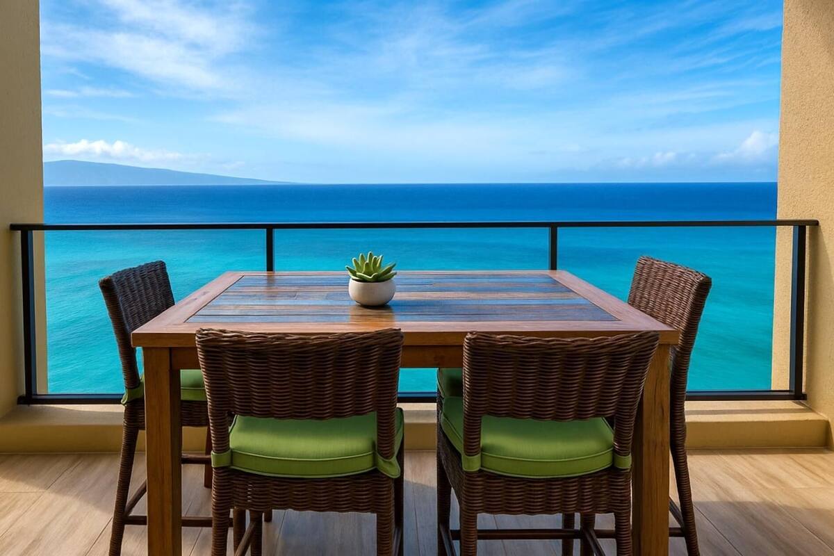 A balcony with a wooden table and wicker chairs, overlooking a clear blue sea and distant mountains.