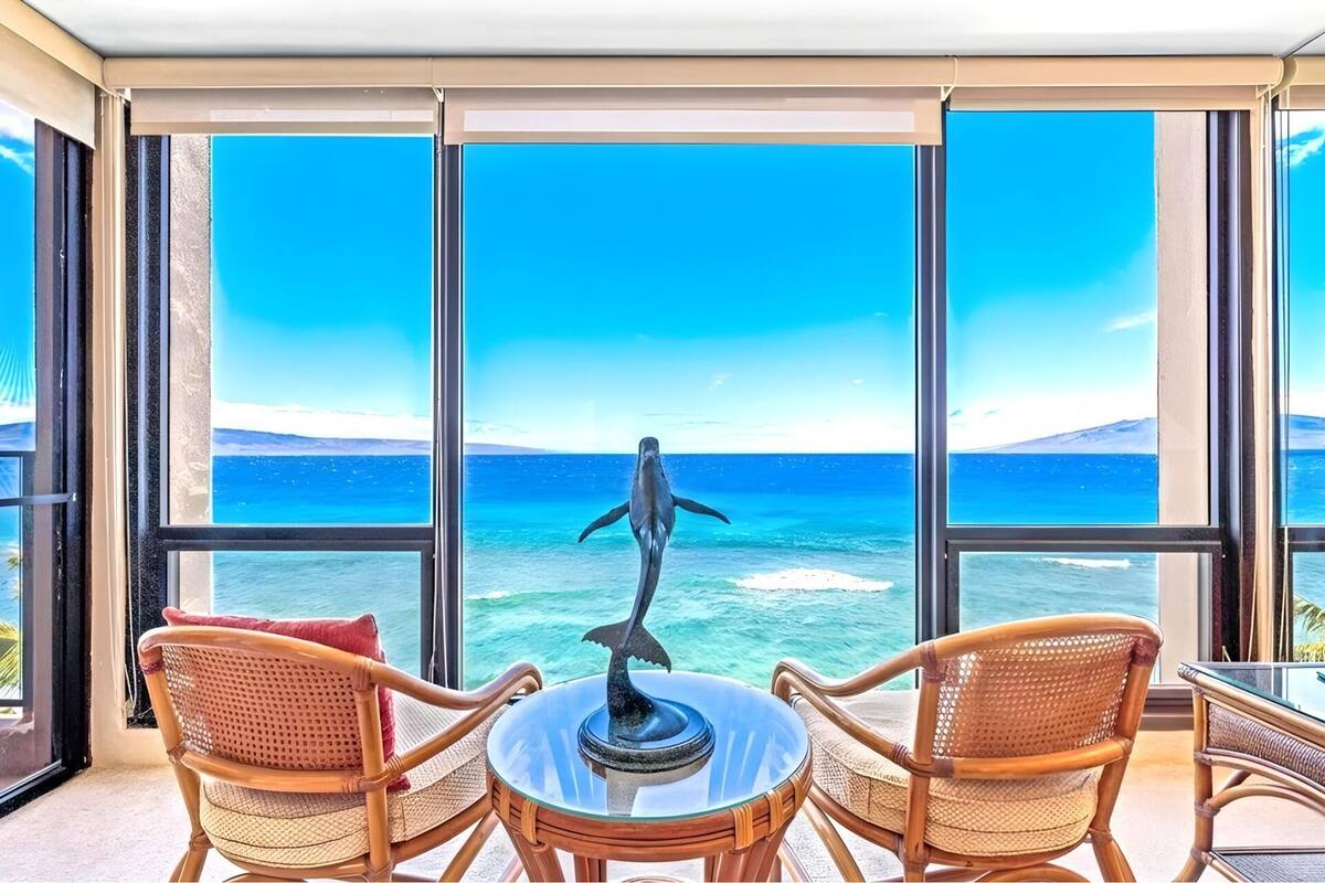 A balcony with wicker chairs and a table, overlooking a clear blue ocean with a whale sculpture.