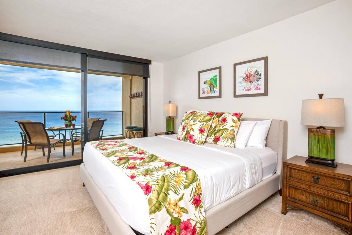 A bedroom with a bed, a nightstand, a chair, a table, and a view of the ocean.