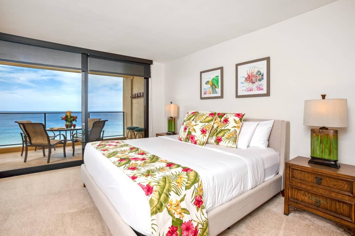 A hotel room with a bed, a nightstand, a chair, a table, and a view of the ocean.