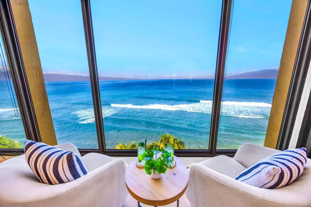A room with a large window overlooking the ocean, two chairs, a small table with a plant, and a striped pillow.