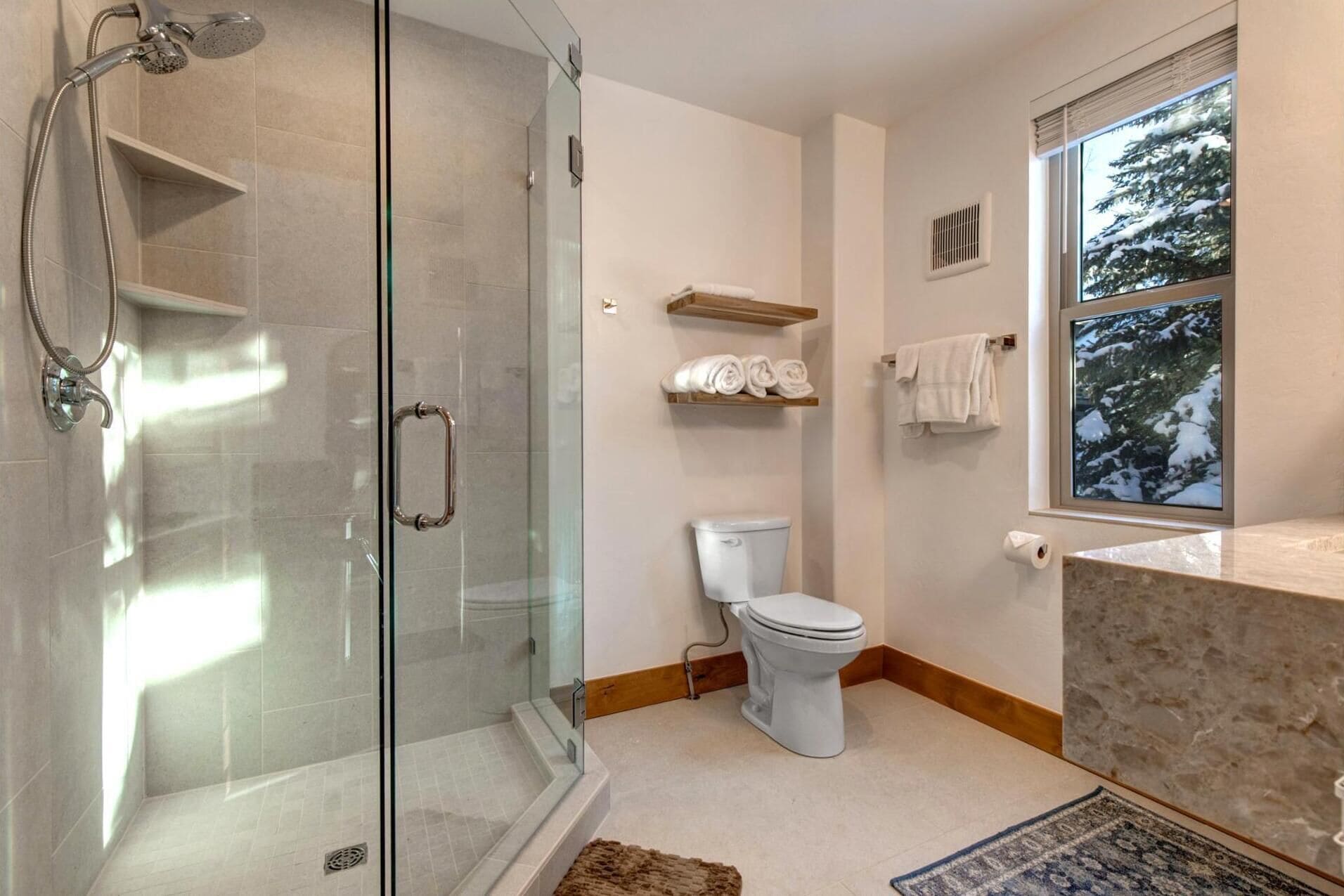 Un baño moderno con ducha envuelta en vidrio, inodoro, lavamanos con encimera de mármol y ventana con vistas a árboles cubiertos de nieve.