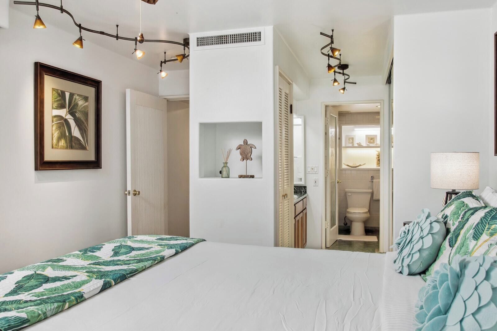 A bedroom with a bed, a nightstand, a lamp, a framed picture, and a hallway leading to a bathroom.