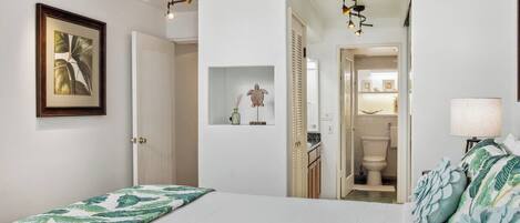 A bedroom with a bed, a nightstand, a lamp, a framed picture, and a hallway leading to a bathroom.