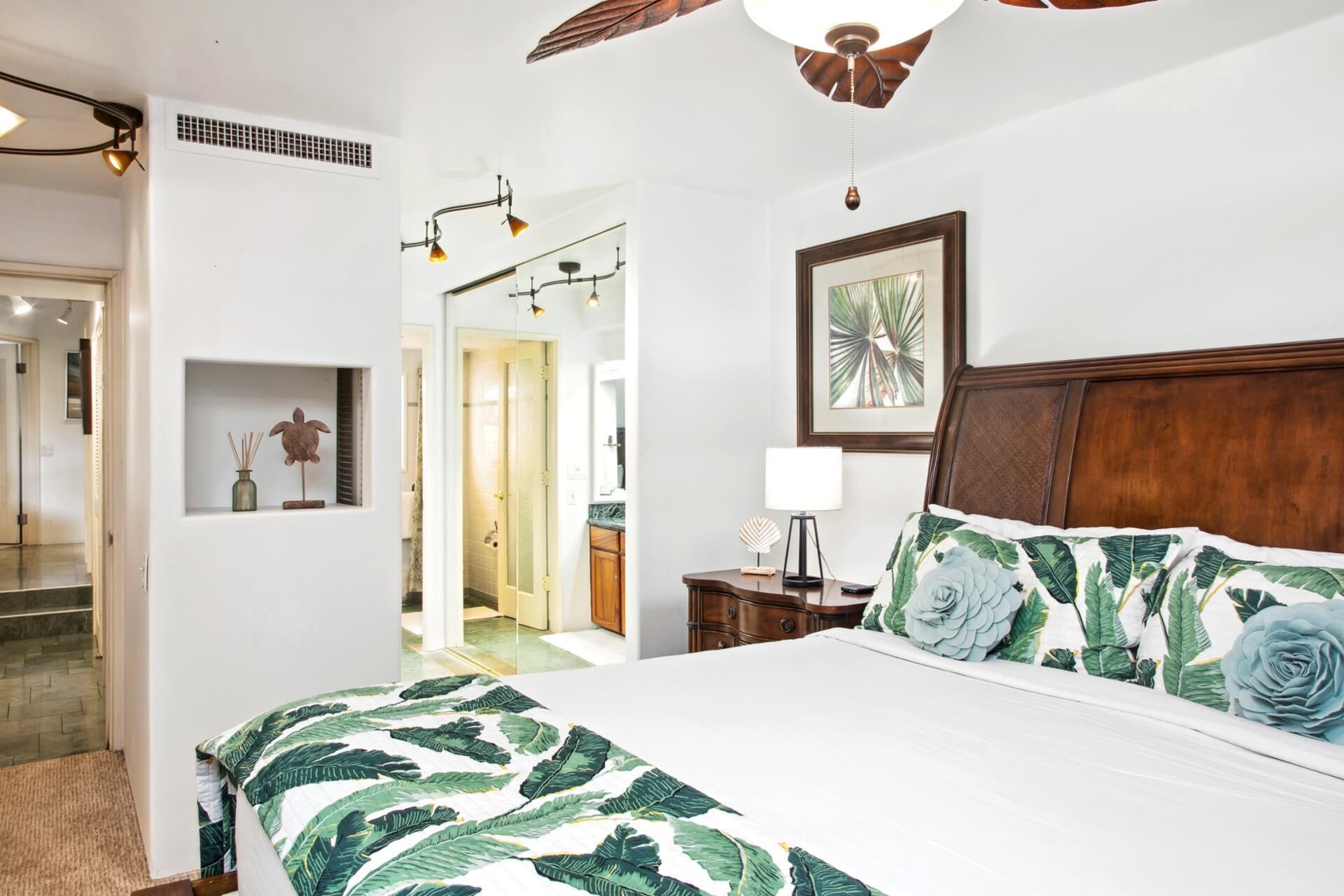 A bedroom with a large bed, a wooden headboard, a ceiling fan, a framed picture, and a nightstand with a lamp.