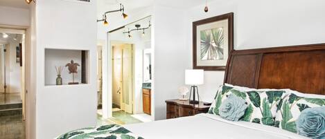 A bedroom with a large bed, a wooden headboard, a ceiling fan, a framed picture, and a nightstand with a lamp.
