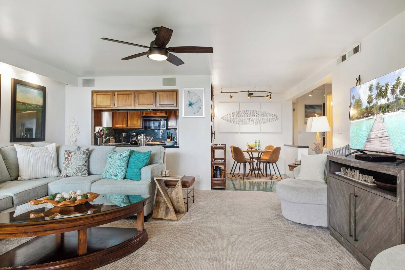 A spacious living room with a sofa, coffee table, and TV stand, a dining area with a table and chairs, and a kitchen with wooden cabinets and a ceiling fan.