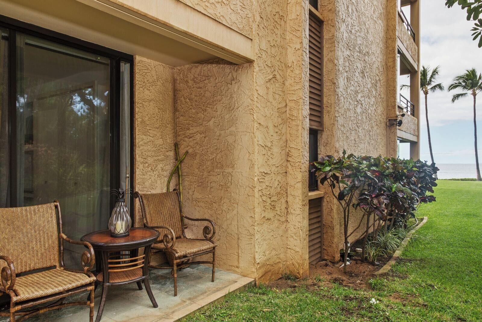 A patio with wicker furniture and a vase, a building with textured walls, and a view of the ocean.