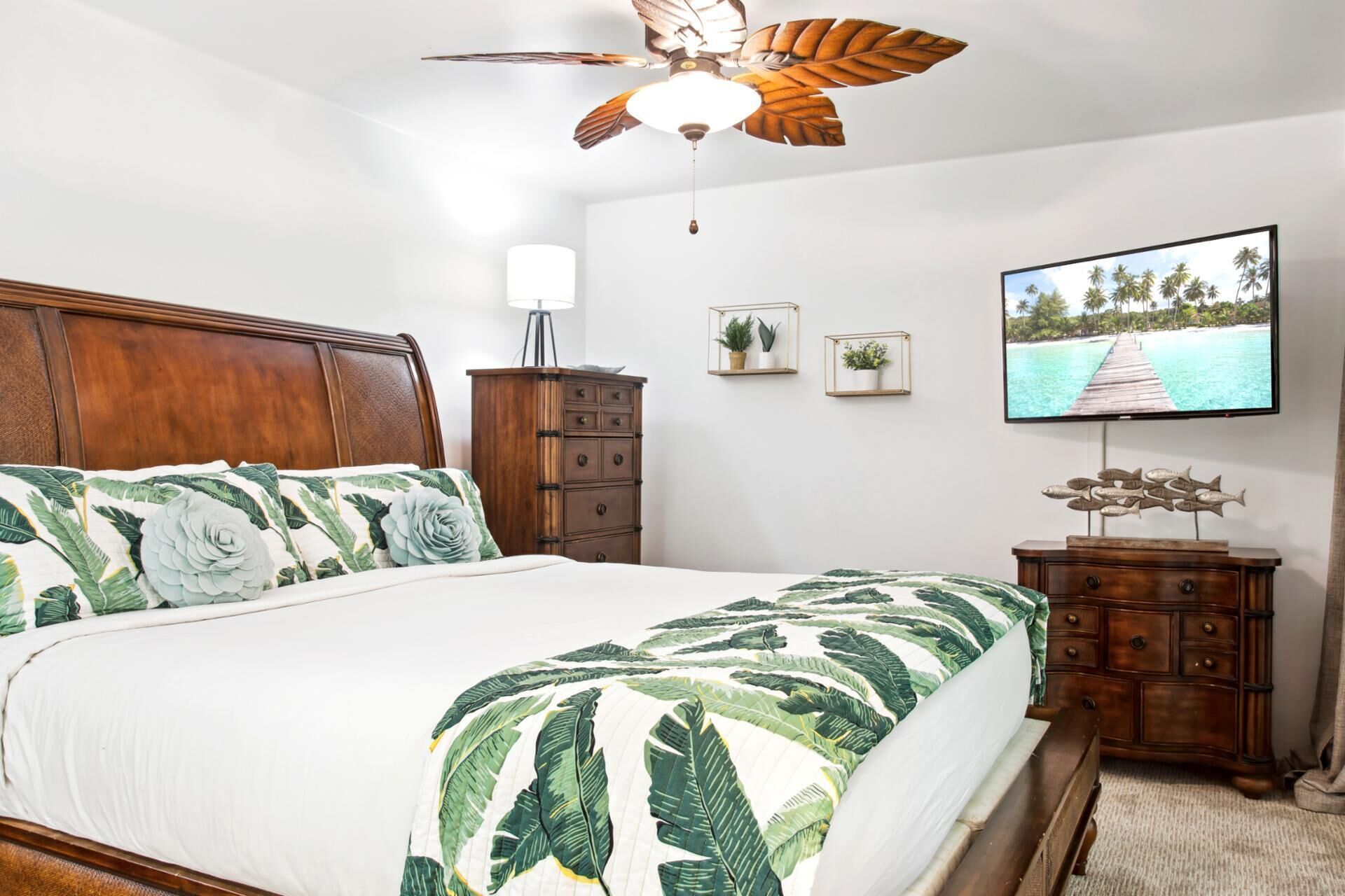 Quarto com cama, cabeceira de madeira, ventoinha no teto, cômoda, TV fixada na parede e quadros emoldurados.