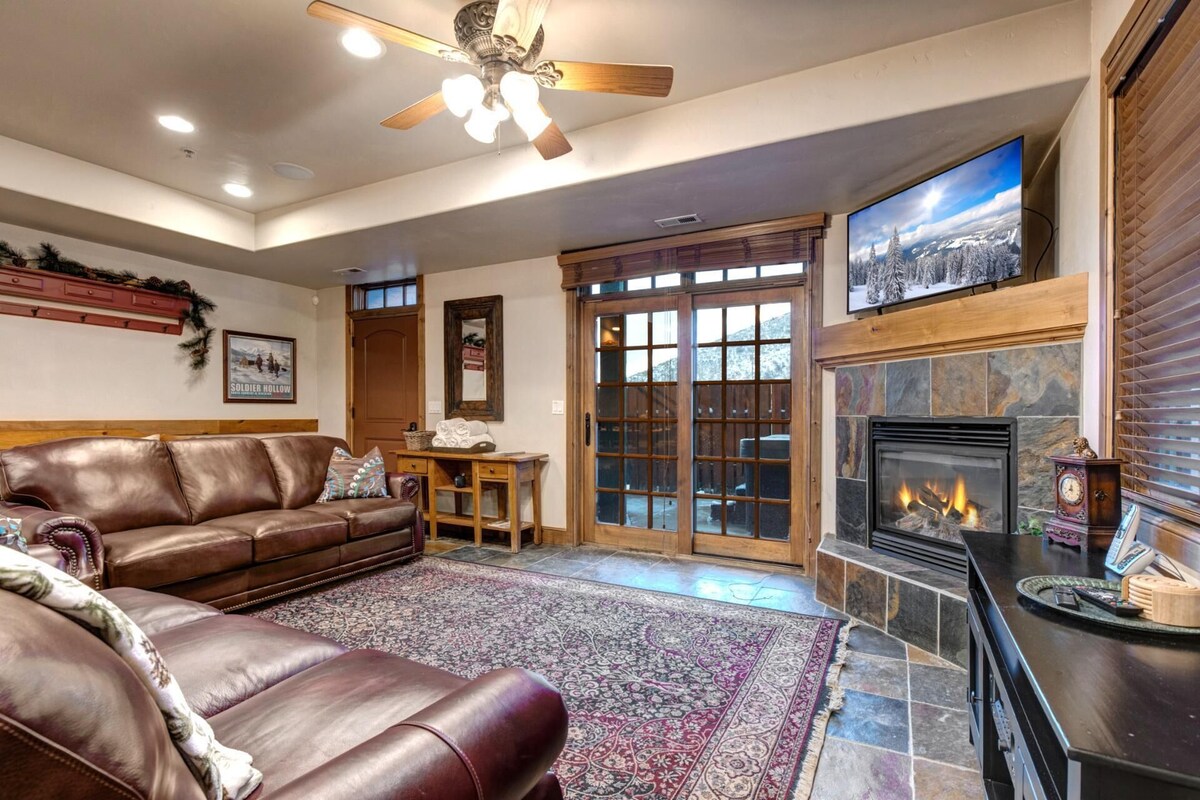 A cozy living room with a leather sofa, a fireplace, and a large window with a view of snow-covered trees.