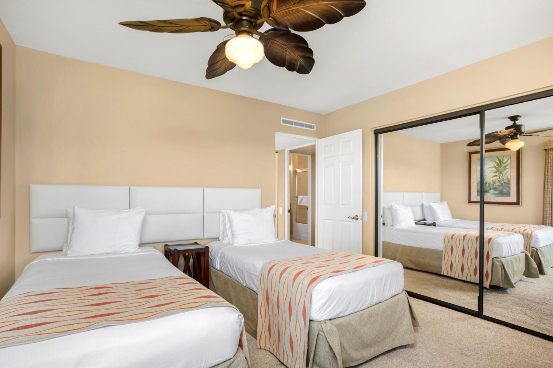 Habitación de hotel con dos camas, un ventilador de techo, un espejo grande y una puerta que da a un baño.