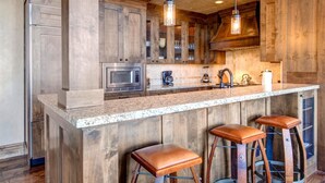 A kitchen with wooden cabinets, a granite countertop, a built-in microwave, a coffee maker, and two bar stools. - Vintage on the Strand by KBM (Park City)