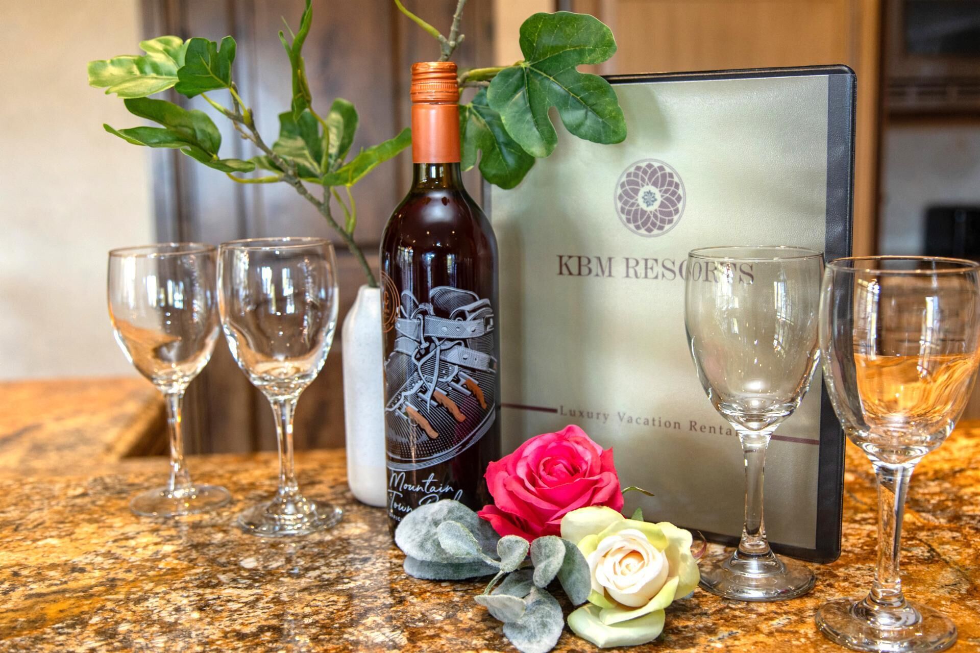 A bottle of wine, two empty wine glasses, a bouquet of roses, and a luxury vacation rental sign.