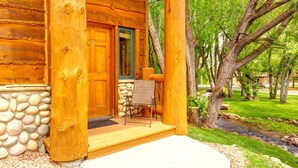 A wooden cabin with a stone foundation, a porch with a chair, and a view of a green landscape with trees and a stream. - Timber Wolf Lodge by KBM (Park City)