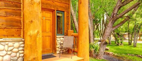 Una cabaña de madera con base de piedra, un porche con una silla y vistas a un paisaje verde con árboles y un arroyo.