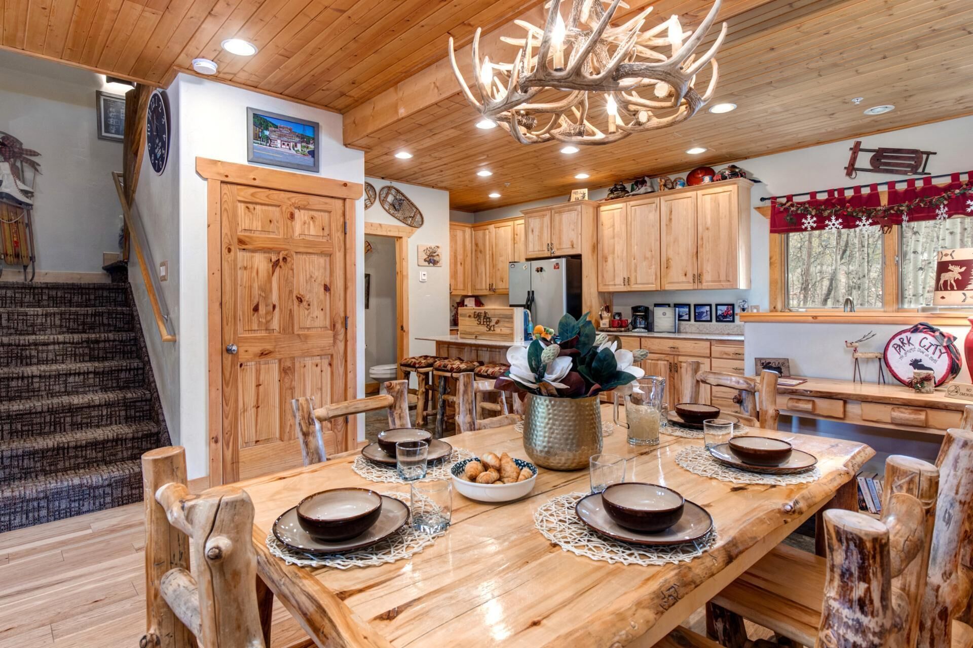 A wooden dining table set with plates and bowls, surrounded by rustic chairs. A chandelier hangs from the ceiling, and wooden cabinets line the walls.