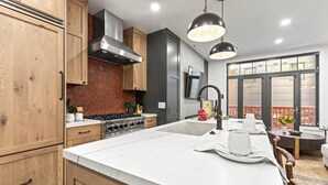 Una cocina moderna con armarios de madera, estufa de acero inoxidable, una isla grande con fregadero, iluminación colgante y vistas a la ciudad a través de amplios ventanales.