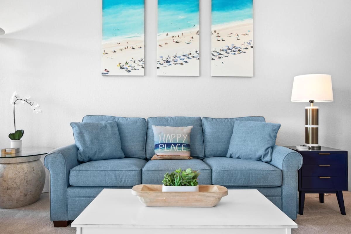 A living room with a blue sofa, a white coffee table, a side table with a lamp, and a wall with three beach-themed paintings.