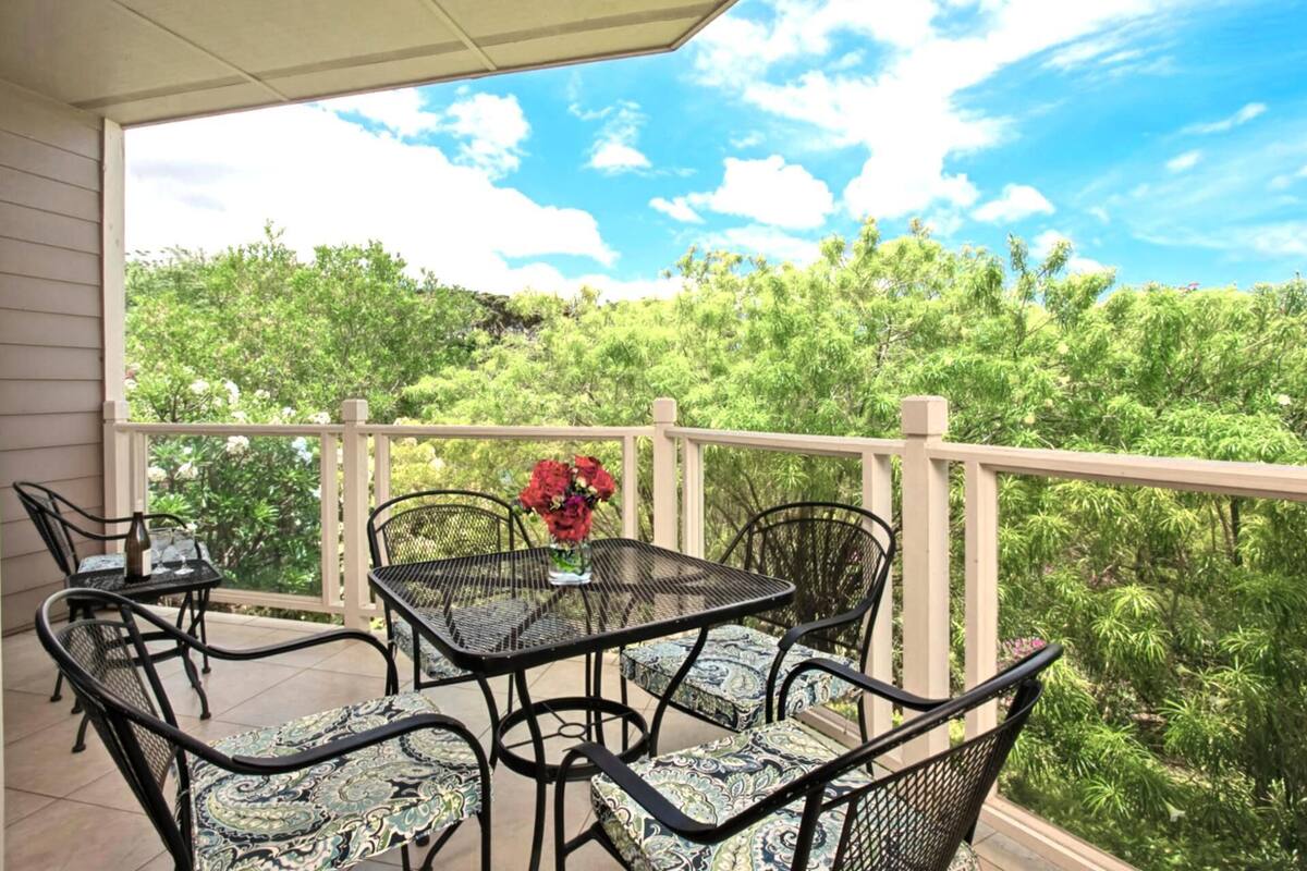 A covered outdoor patio with a table and chairs, surrounded by a white railing, overlooking a lush green landscape.