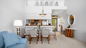 A modern living room with a blue sofa, a dining area with white chairs, a kitchen with stainless steel appliances, and a large 'ALOHA' sign above the kitchen. - Grand Champions by KBM (Kihei)