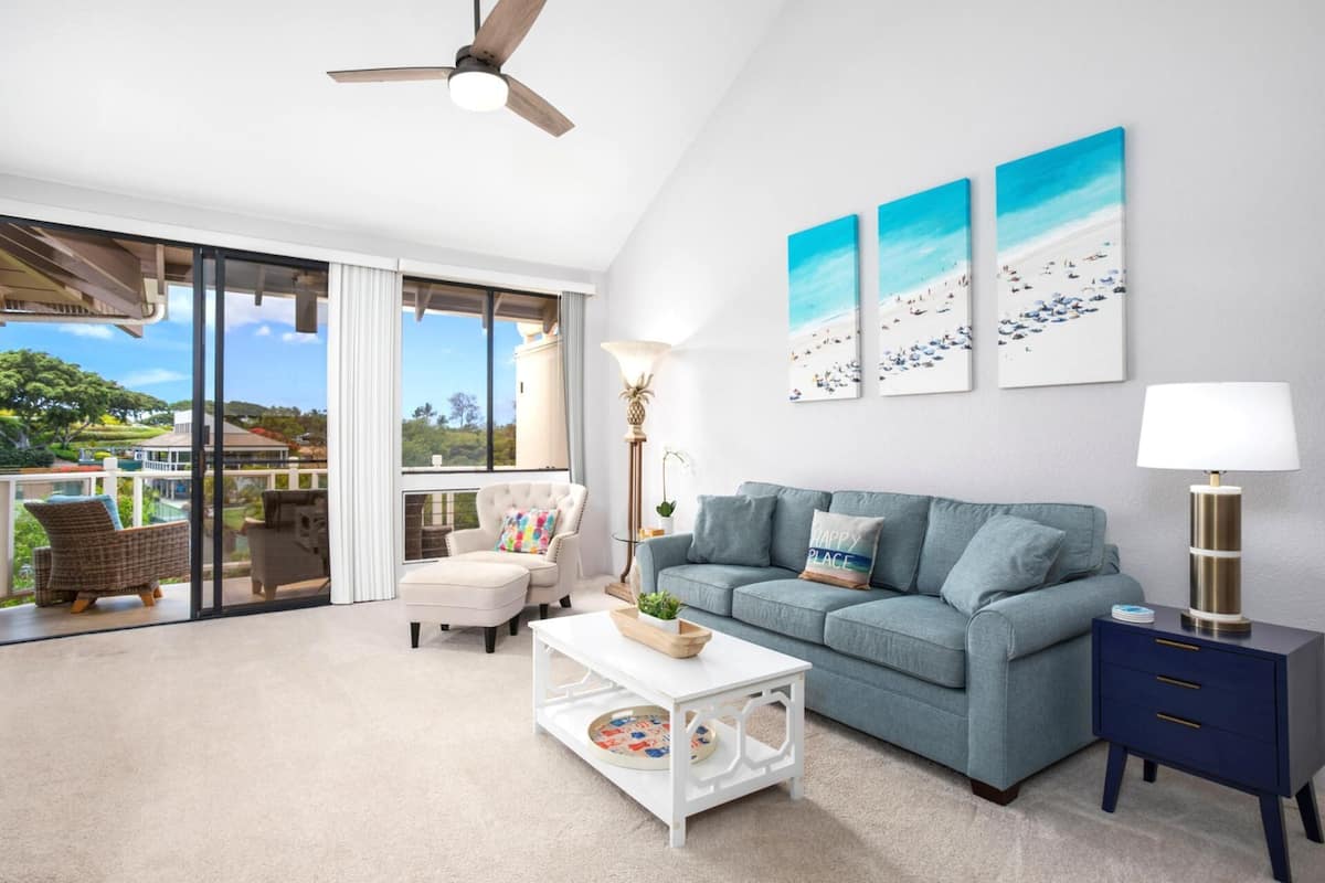 A spacious living room with a large sliding glass door leading to a balcony, a light blue sofa, a white coffee table, and a blue side table.