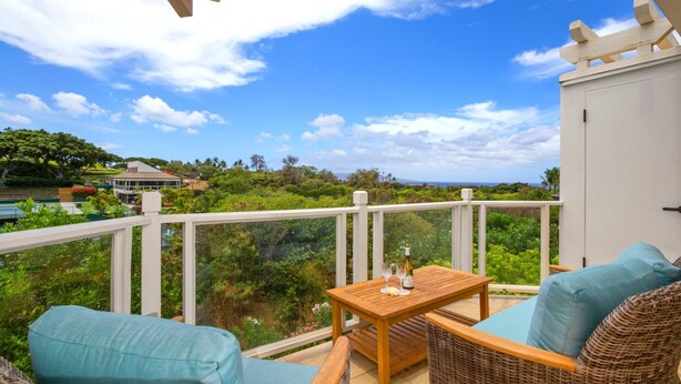 A balcony with wicker furniture, a wooden table, and a view of a lush landscape and distant buildings. - Grand Champions by KBM (Kihei)