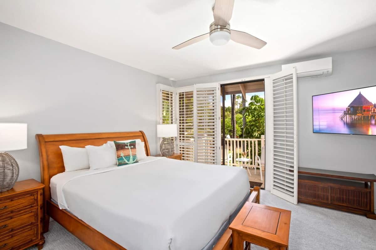 A bedroom with a bed, bedside tables, a ceiling fan, a TV, and a balcony with shutters.