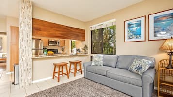 Uma sala de estar moderna com um sofá cinza, dois banquinhos de madeira, um tapete estampado e uma cozinha com armários de madeira e eletrodomésticos de aço inoxidável.
