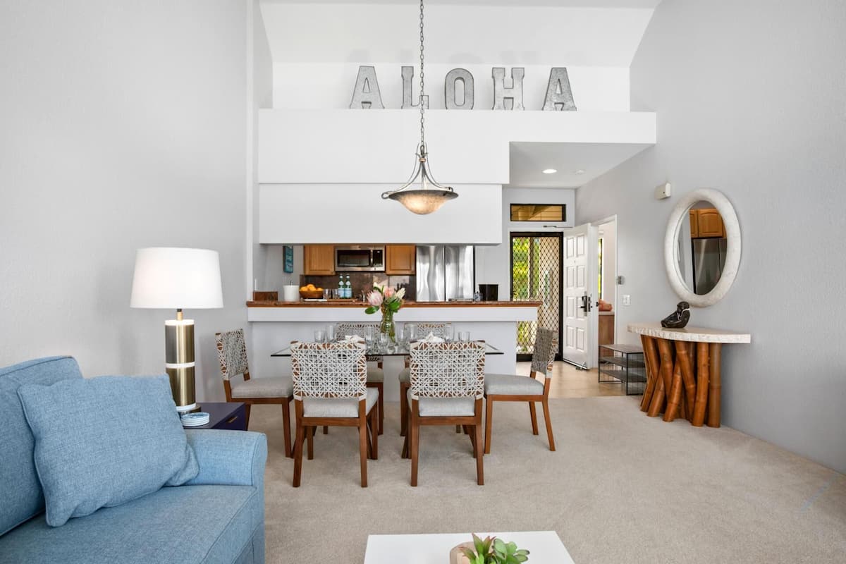 A modern living room with a blue sofa, a dining area with white chairs, a kitchen with stainless steel appliances, and a large 'ALOHA' sign above the kitchen.