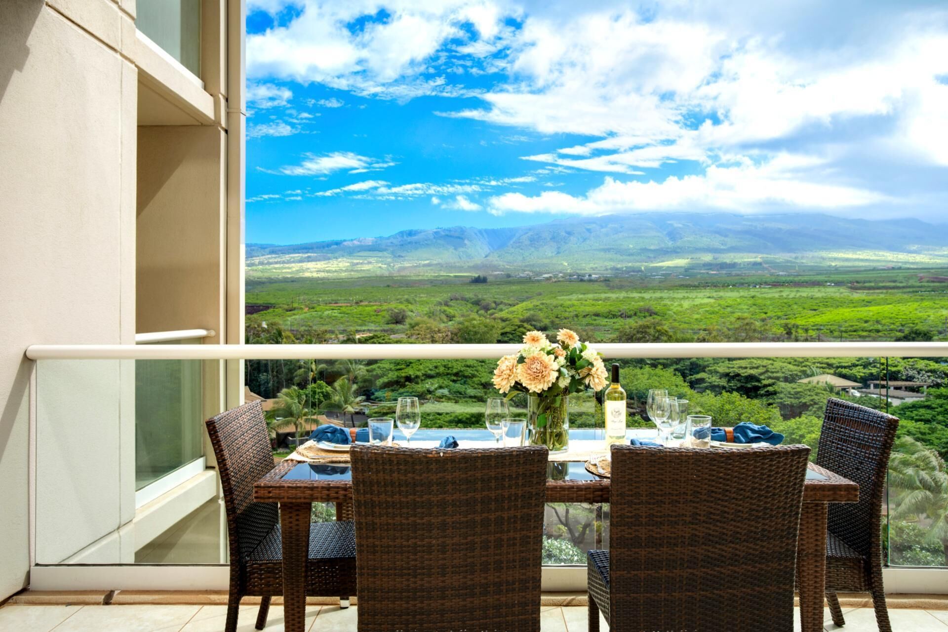 A dining table with wicker chairs and a vase of flowers, set on a balcony with a scenic view of green hills and mountains.