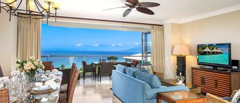 A hotel room with a dining area, a sofa, a TV, and a view of the ocean.