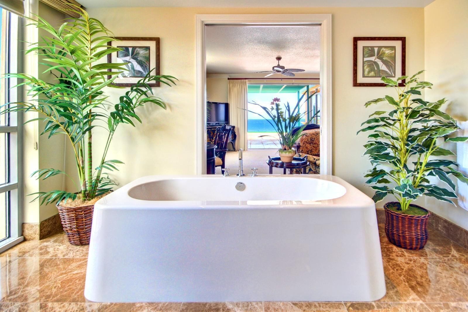 A bathroom with a large bathtub, two potted plants, and a view of the beach through a doorway.