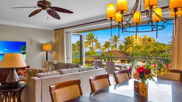 A hotel room with a balcony, a ceiling fan, a TV, a dining area with chairs and a table, and a view of palm trees and buildings.