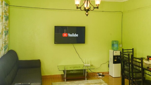 A living room with a flat-screen TV mounted on the wall, a chandelier, a sofa, a small table, and a water dispenser. - Stay.Plus Lake View House Naivasha (Naivasha)