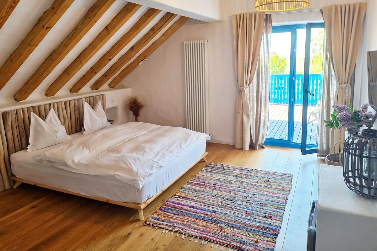 A bedroom with a wooden ceiling, a bed with white bedding, a colorful woven rug, and a view of the outside.