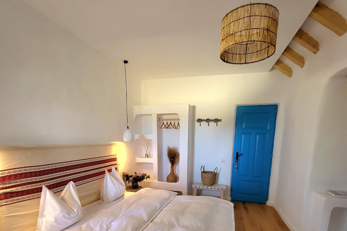 A bedroom with a bed, a blue door, a hanging light fixture, and a wall-mounted shelf with decorative items.