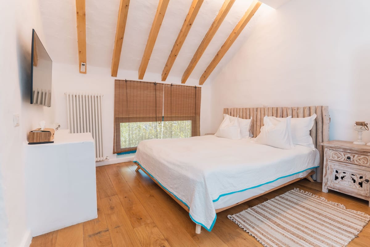A bedroom with a large bed, wooden beams on the ceiling, a radiator, a window with blinds, a bedside table, and a TV mounted on the wall.