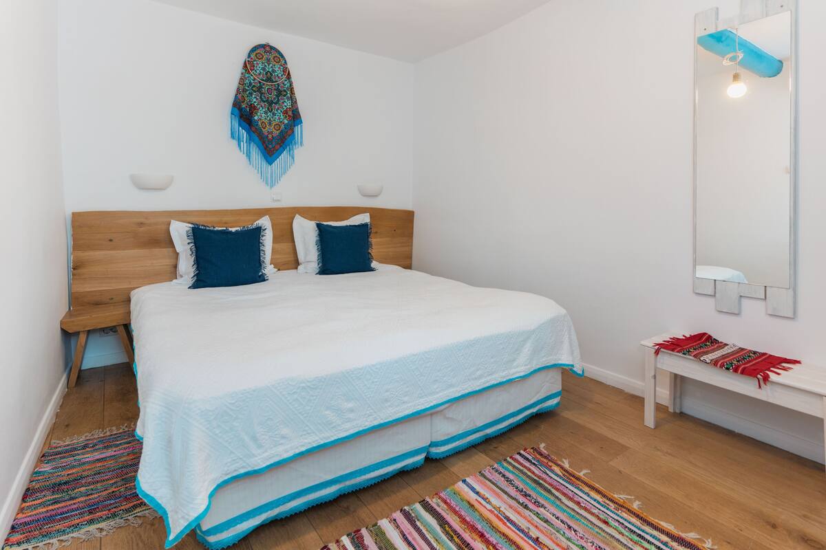 A bedroom with a wooden headboard, a white bedspread, two blue pillows, a colorful woven rug, and a white bench with a red and green woven blanket.