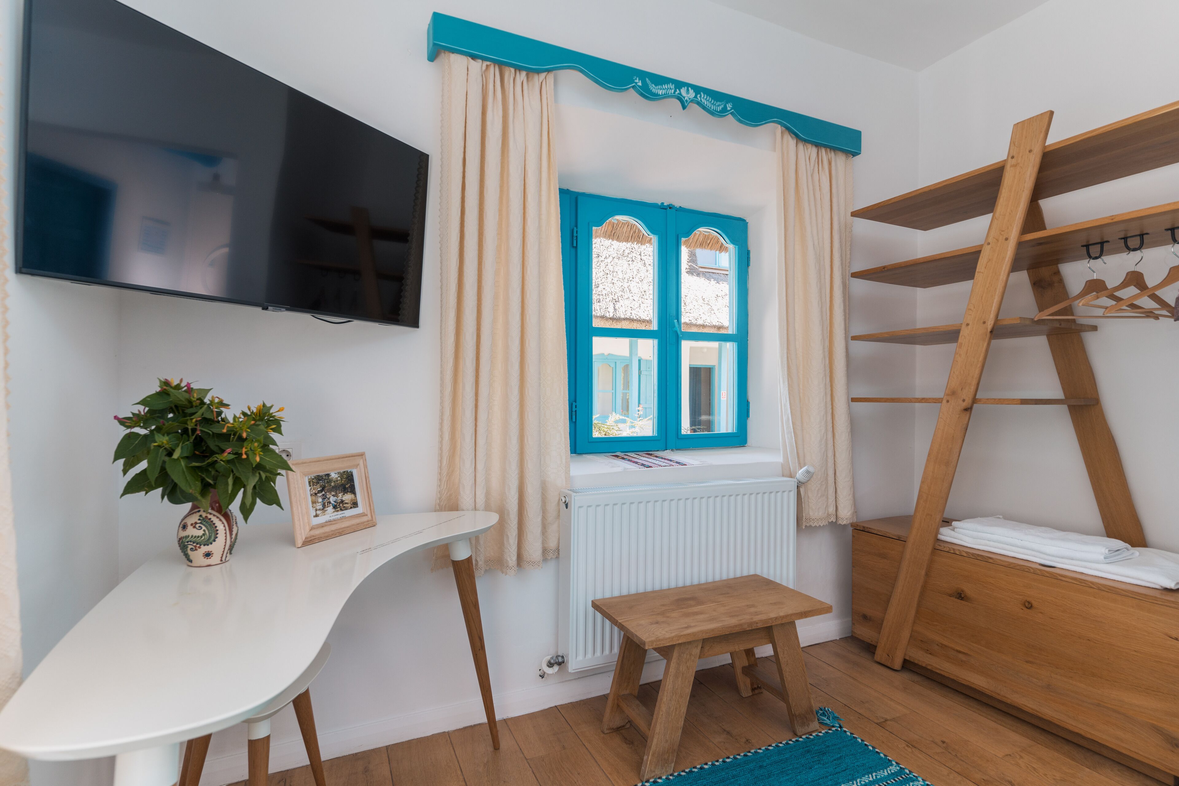 Una habitación con televisión montada en la pared, ventana con contraventanas azules, estante de madera, una mesita con una planta y un banco.