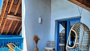 Una terraza de madera con muebles de mimbre, incluyendo sillas y una mecedora colgante, frente a un muro azul con un ventanal de vidrio.